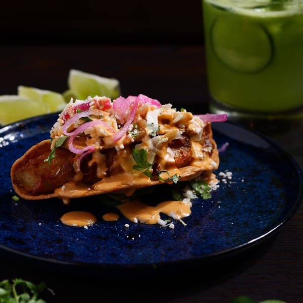 A Str8 up Tacos fish taco, topped with pink pickled onions, sauce, and cheese, on a blue plate.