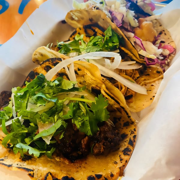 Two Str8 up Tacos, one with meat and cilantro, the other with slaw and sauce, in a white paper tray.